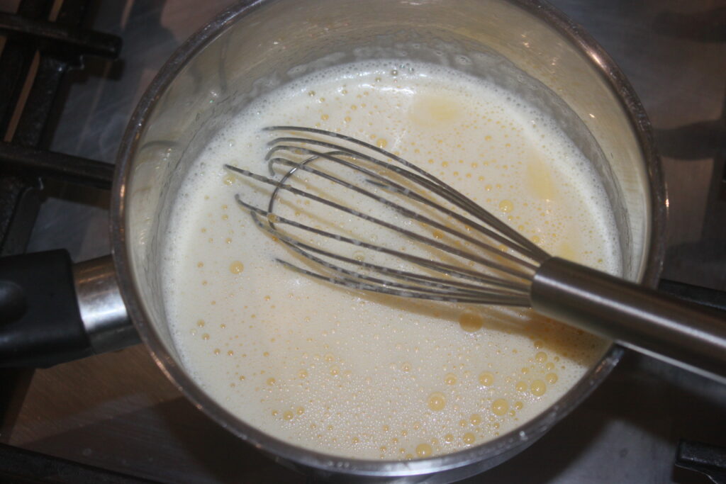 Un fouet en métal remue un liquide crémeux et pétillant dans une casserole en acier inoxydable sur la cuisinière, créant ainsi les Tartelettes à la crème d'orange parfaites.