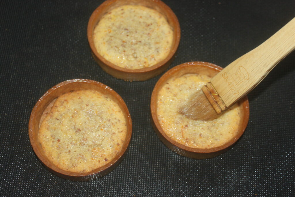 Trois tartelettes à la crème d'orange, garnies d'un mélange crémeux, sont posées sur une surface sombre. Un pinceau applique le glaçage sur les desserts, mettant en valeur les saveurs subtiles d'agrumes sous leur extérieur doré.