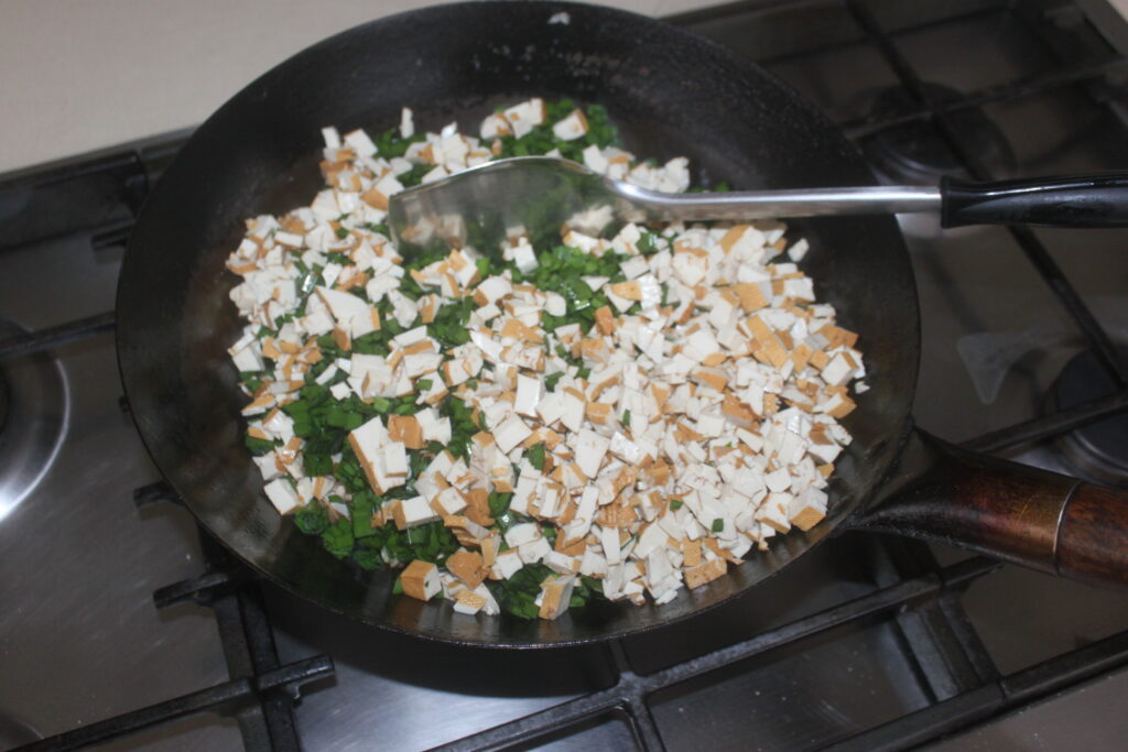 Du tofu coupé en dés, sauté avec des champignons shiitake et des oignons verts hachés, grésille dans une poêle sur la cuisinière, à côté d'une spatule fiable.