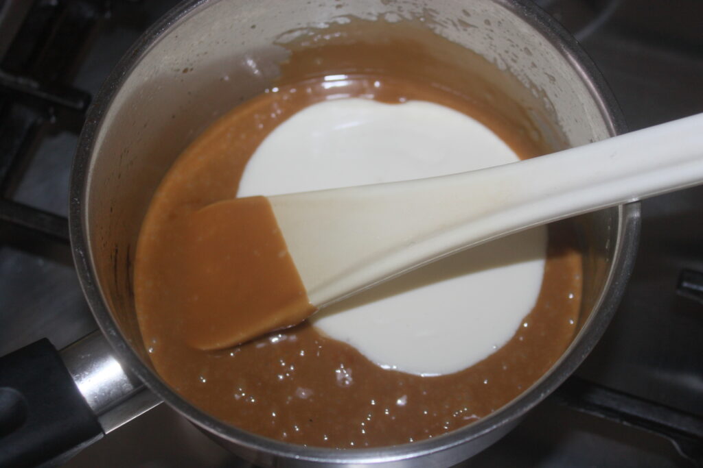 Une casserole sur la cuisinière contient du caramel fondu et de la crème, savamment mélangés avec une spatule blanche, créant une base succulente qui suggère une version ludique de la traditionnelle Galette des Rois.