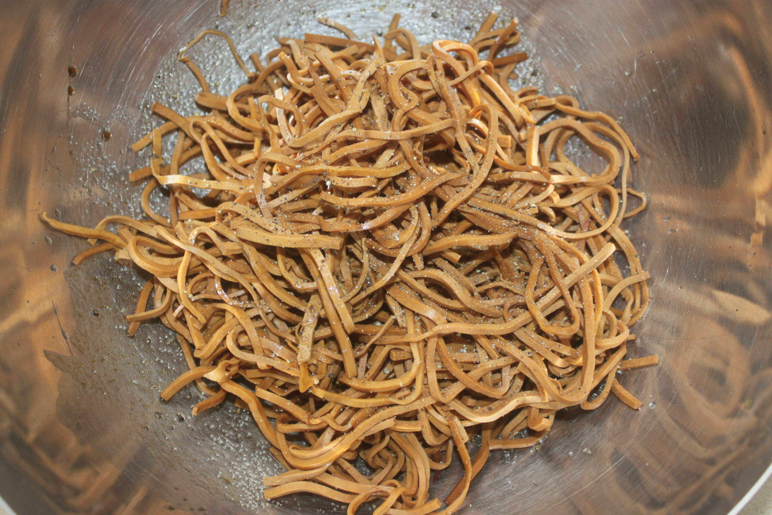 Des nouilles brunes cuites assaisonnées de poivre noir sont servies dans un bol en métal, capturant l'essence d'un repas auto Brouillon.