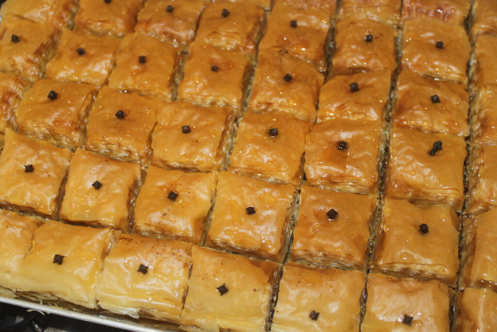 Un plateau de carrés de baklava brun doré, chacun surmonté d'un seul clou de girofle et parsemé de noix finement hachées, présente un délicieux mélange de textures et de saveurs inspirées de la cuisine grecque traditionnelle.
