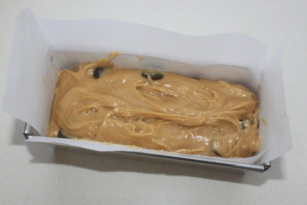 Un moule à pain tapissé de papier sulfurisé est rempli d'une irrésistible pâte à la cacahuète et à la banane, parsemée de pépites de chocolat, à laquelle il est presque impossible de résister avant la cuisson.