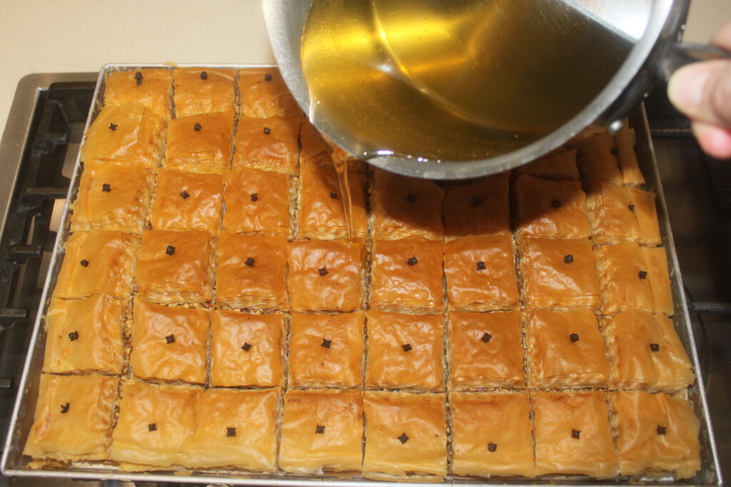 Un plateau de carrés de baklava arrosés de sirop provenant d'un pot, chacun orné d'un clou de girofle au centre. Ces friandises grecques regorgent de couches de pâtisserie délicate et de noix croquantes, créant une symphonie de saveurs à chaque bouchée.