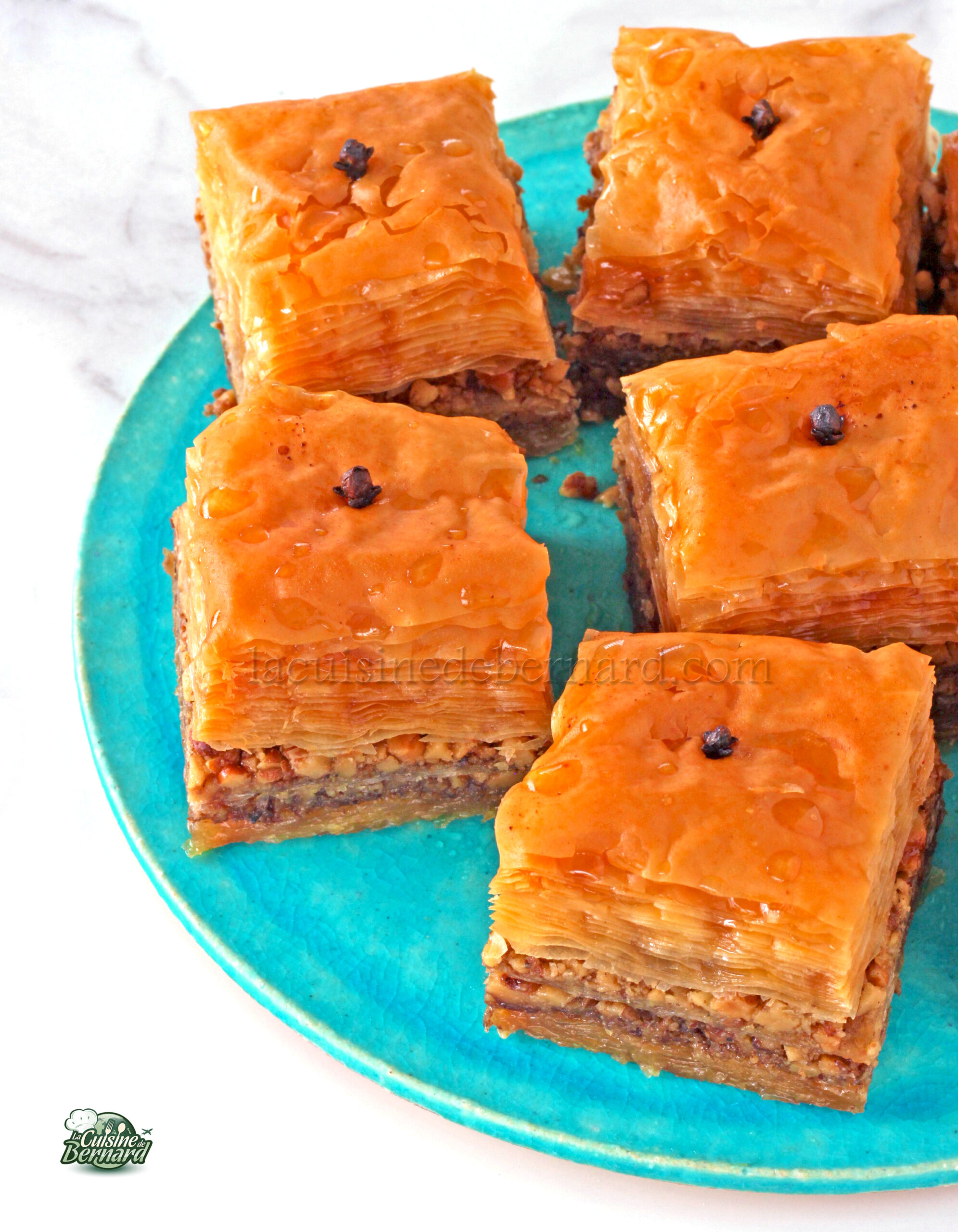 Cinq morceaux de baklava, une pâte feuilletée fourrée aux noix et sucrée au sirop, sont disposés sur une assiette turquoise.