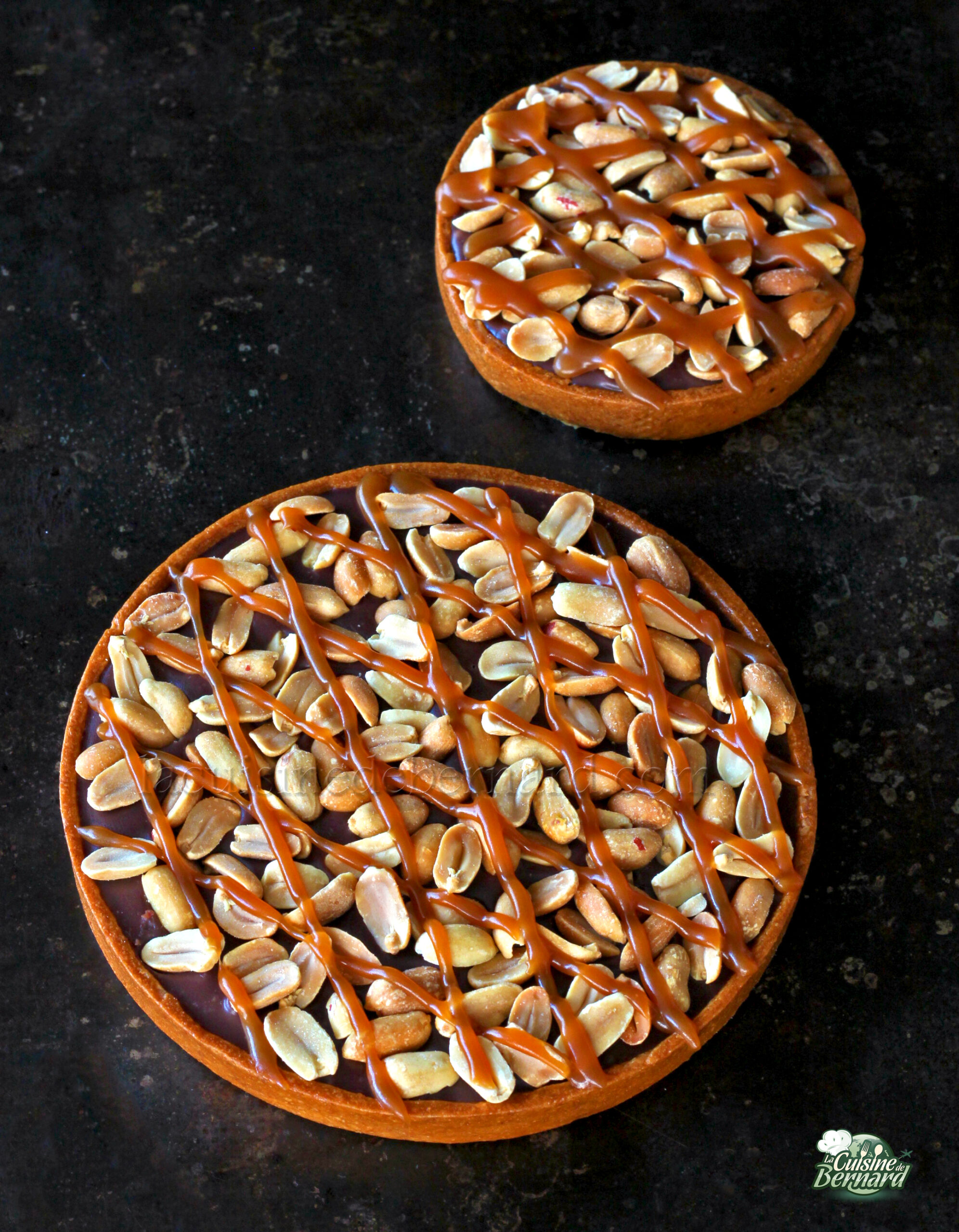 Deux tartes dessert rondes, garnies de cacahuètes et arrosées de caramel, posées sur une surface sombre.