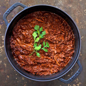 Un pot de viande effilochée dans une sauce riche et foncée, garnie d'un brin de persil, placé sur une surface sombre.